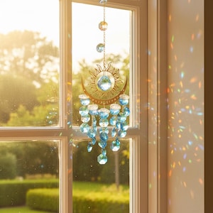 May include: A decorative suncatcher hanging in front of a window. The suncatcher features a gold-coloured sun and crescent moon design, with hanging blue crystal beads. The sunlight creates a rainbow effect on the wall.