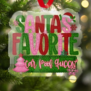 以下が含まれることがあります： 緑、赤、ピンクで「SANTA'S FAVORITE car pool QUEEN」と書かれた透明なクリスマスオーナメント。オーナメントには吊り下げ用の赤いリボンが付いており、クリスマスツリー、雪の結晶、サンタの帽子が描かれています。