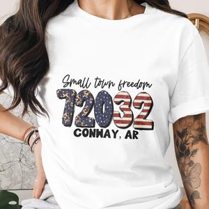 Puede incluir: Camiseta blanca con el texto "Small town freedom" y los números "12032" en un diseño patriótico. El texto "CONWAY, AR" está impreso debajo. La camiseta es de estilo casual.