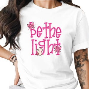 May include: White t-shirt with the pink text "Be the Light", a bunny, and bows. The design is on the chest. A casual, everyday tee.