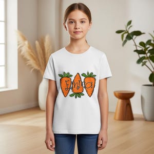 Op de afbeelding: Wit T-shirt met drie oranje wortel illustraties met groene toppen en de naam "Emma" in zwart script. De wortels hebben zwarte contouren. Het T-shirt wordt door een persoon gedragen.