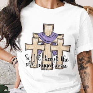 May include: White t-shirt featuring a graphic of three wooden crosses with a purple cloth draped over them. The text "So I'll cherish the old rugged cross" is printed below the crosses in a script font.