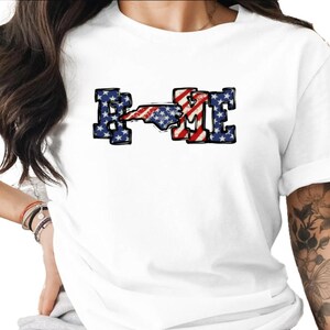 Puede incluir: Camiseta blanca con la palabra "HOME" en un diseño patriótico. Las letras "H" y "E" son azules con estrellas blancas, y la "O" y el contorno de Carolina del Norte son de rayas rojas y blancas.