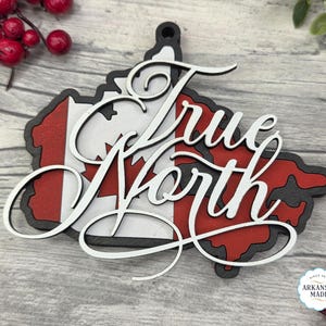 May include: A wooden ornament in the shape of Canada, featuring the Canadian flag colors and the words "True North" in elegant script. The ornament has a black border and a hole for hanging. The ornament is made in Arkansas.