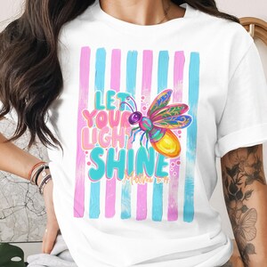 May include: White t-shirt with a colourful firefly illustration and the text "Let Your Light Shine." The design incorporates pink and blue vertical stripes. The firefly is rendered in bright, eye-catching colours.