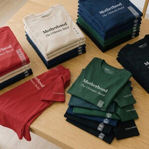 May include: Stacks of t-shirts in various colors, including red, beige, navy, green, and black, are arranged on a wooden surface. Each shirt features the text "Motherhood The Ultimate Build" in white. Sizes XS to 4XL are visible.