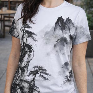 May include: A white short-sleeved t-shirt featuring a black and gray ink wash style landscape print. The graphic depicts jagged mountain peaks, misty valleys, and detailed pine trees extending across the front and onto the sleeves.
