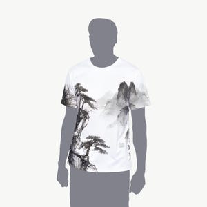 May include: A white short-sleeve t-shirt featuring a black and gray ink wash style landscape print. The graphic depicts detailed pine trees growing on rocky cliffs with misty mountains in the background, covering the front of the garment.