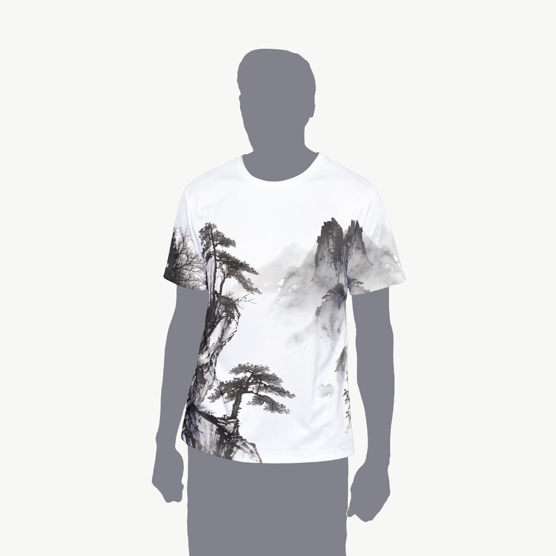 May include: A white short-sleeve t-shirt featuring a black and gray ink wash style landscape print. The graphic depicts detailed pine trees growing on rocky cliffs with misty mountains in the background, covering the front of the garment.