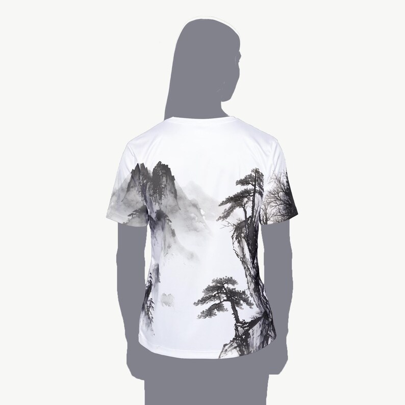 May include: A white short-sleeve t-shirt featuring a black and gray ink wash style landscape print. The graphic depicts jagged mountain peaks, rocky cliffs, and pine trees in a traditional artistic style that wraps around the back of the garment.