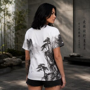 May include: A white short-sleeve t-shirt featuring a black and gray ink wash style landscape print on the back. The graphic depicts detailed mountain peaks, rocky cliffs, and pine trees in a traditional artistic style. The shirt has a crew neck.