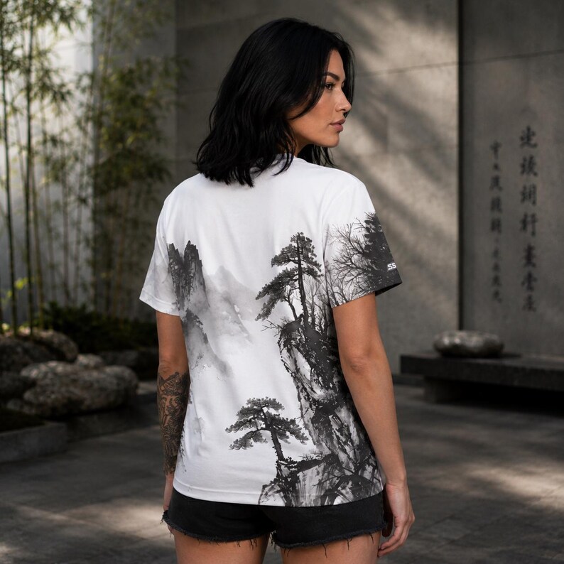 May include: A white short-sleeve t-shirt featuring a black and gray ink wash style landscape print on the back. The graphic depicts detailed mountain peaks, rocky cliffs, and pine trees in a traditional artistic style. The shirt has a crew neck.