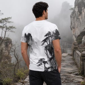 May include: A white short-sleeve t-shirt featuring a black and grey ink wash style landscape print. The graphic depicts misty mountains and pine trees extending across the back and side. The shirt has a crew neckline and a small logo on the sleeve.