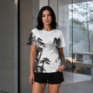 May include: A white short-sleeve t-shirt featuring a black and gray ink wash style landscape print. The graphic depicts jagged mountain peaks, rocky cliffs, and pine trees in a traditional East Asian art style across the front and sleeve.