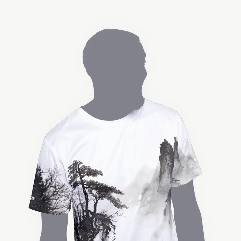 May include: A white short-sleeve t-shirt featuring a black and gray ink wash style graphic print. The design depicts a detailed landscape with tall pine trees and jagged mountain peaks, extending across the front and onto the right sleeve.
