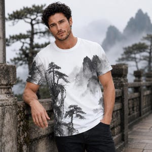 May include: A white short-sleeve t-shirt featuring a black and gray ink wash style landscape print. The graphic depicts jagged mountain peaks, pine trees, and misty valleys, extending across the front and onto the sleeve of the garment.