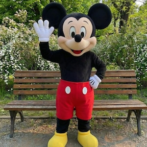 May include: A person in a Mickey Mouse costume, waving. The costume features a black top, red shorts with white buttons, yellow shoes, and white gloves. The background includes a wooden bench and greenery.