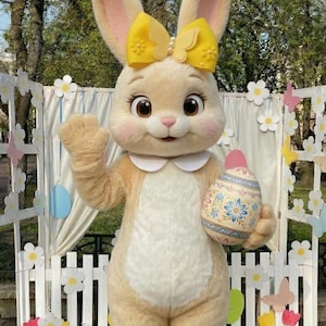 Può includere: Una persona in un costume da coniglio marrone chiaro con un fiocco giallo che tiene in mano un uovo di Pasqua decorato. Il coniglio è in piedi davanti a una staccionata bianca decorata con fiori e farfalle. Il costume ha un colletto bianco.
