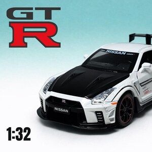 May include: A white and black Nissan GT-R model car with a rear spoiler. The model has black rims with red accents and the text "NISSAN" on the windshield. The image also includes the text "GT R" and "1:32".