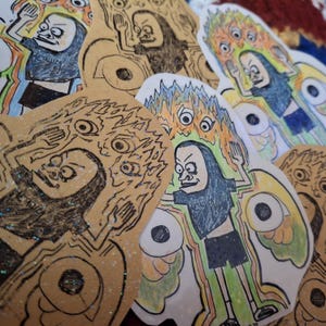 Op de afbeelding: Een verzameling kleurrijke, handgetekende stickers met gestileerde figuren met overdreven kenmerken. De illustraties bevatten figuren met meerdere ogen, vlammen en een verscheidenheid aan kleuren, waaronder zwart, bruin, blauw, groen en oranje.