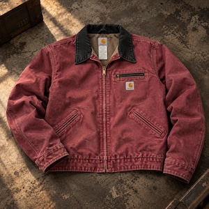 Puede incluir: Una chaqueta de lona roja con cuello de pana negro, cierre de cremallera frontal y forro de forro polar beige. La prenda incluye un bolsillo en el pecho con cremallera, dos bolsillos inferiores y un parche de la marca Carhartt en el pecho.