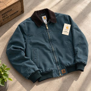 May include: A teal Carhartt jacket with a dark brown corduroy collar, a full-length brass zip, and ribbed cuffs and hem. The jacket features a western-style yoke, two front pockets, and a white brand tag attached to the chest with the text RUGGED OUTDOOR WEAR.