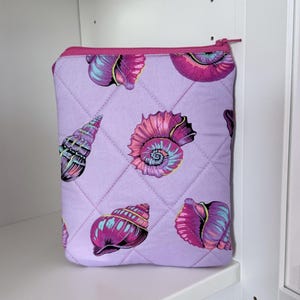 May include: A quilted fabric pouch in a light lavender color featuring a repeating pattern of seashells in shades of magenta, purple, and teal. The rectangular pouch has a matching magenta zipper closure along the top edge and diamond stitching.