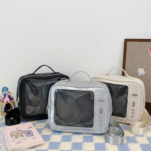 Può includere: Tre borse quadrate a forma di televisori vintage. Le borse sono nere, argentate e color crema, ognuna con uno schermo trasparente e un manico. Sono esposte su una superficie a scacchi blu e bianca.