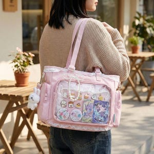 May include: A pink tote bag with a clear front pocket, decorated with lace trim and a white bunny charm. The bag features a variety of colourful pins and charms inside the clear pocket, and a pearl necklace. The bag has a shoulder strap and two handles.