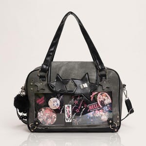 Könnte beinhalten: Graue Tragetasche mit schwarzen Griffen und Sternverzierungen. Die Tasche hat transparente Taschen mit farbenfrohen Grafiken und Abzeichen im Anime-Stil. Ein schwarzer Pompon und ein silberner Schultergurt ergänzen das Design.