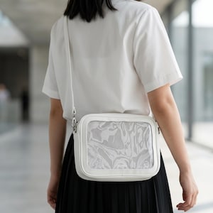 Puede incluir: Un bolso blanco cruzado con forma rectangular y un panel frontal transparente que muestra un patrón abstracto y arremolinado. El bolso tiene una correa larga y delgada y herrajes plateados. La persona lleva una camisa blanca y una falda negra.