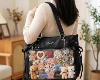 Könnte beinhalten: Schwarze Tragetasche mit einem transparenten Frontpanel, das Plüschtiere, herzförmige Anstecknadeln und dekorative Knöpfe zeigt. Die Tasche hat einen Schulterriemen und das Wort "KAZEVO" ist oben aufgedruckt. Die Worte "DREAM LOVE EXPLORE" sind auf dem transparenten Panel aufgedruckt.