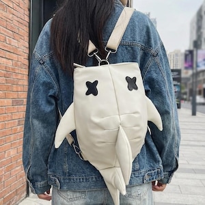 May include: Cream-coloured shark-shaped backpack with black cross-shaped eyes and fins. The backpack is worn by a person wearing a denim jacket and jeans. The bag has adjustable straps and a zip closure.