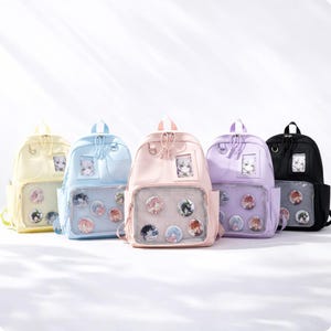 Könnte beinhalten: Fünf pastellfarbene Rucksäcke in Gelb, Hellblau, Rosa, Lila und Schwarz. Jeder Rucksack hat eine transparente Vordertasche mit dekorativen Anstecknadeln und ein kleines Fenster zur Präsentation eines Fotos. Die Rucksäcke haben ein schlichtes, klares Design.