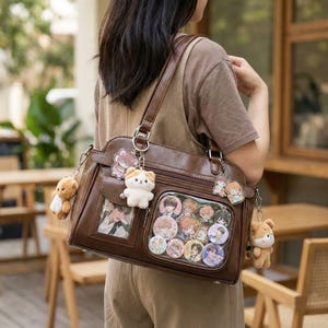 Könnte beinhalten: Eine braune Umhängetasche mit mehreren Fächern und einem transparenten Fenster, das bunte Anstecknadeln zeigt. Die Tasche ist mit verschiedenen Anhängern verziert, darunter kleine Teddybären und Figuren im Anime-Stil. Die Tasche hat einen Schultergurt und einen oberen Griff.