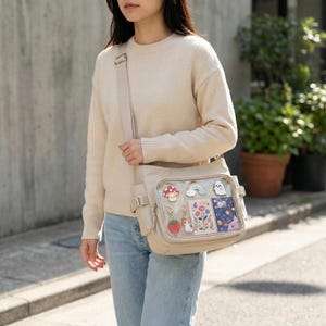 Puede incluir: Bolso bandolera beige con un bolsillo frontal transparente que muestra dijes y tarjetas coloridas. El bolso tiene una correa larga ajustable y múltiples bolsillos. Los dijes incluyen un hongo, un arcoíris, un fantasma, una fresa, un gato y diseños florales.