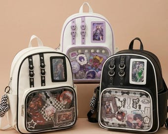 May include: Three backpacks in white, purple, and black. Each has a clear front pocket displaying various decorative items. The white and black backpacks have silver hardware and a small handle. The purple backpack has purple accents.