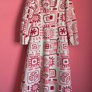 May include: A long-sleeved, floor-length coat featuring a white base color with a repeating pattern of geometric shapes and abstract floral motifs in shades of bright pink and light pink. The coat has a high collar and a structured, straight fit.
