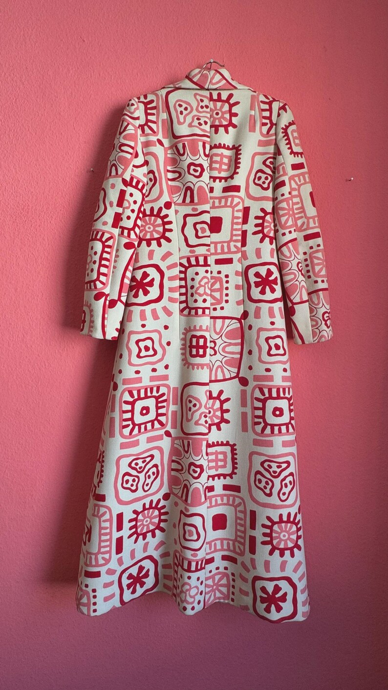May include: A long-sleeved, floor-length coat featuring a white base color with a repeating pattern of geometric shapes and abstract floral motifs in shades of bright pink and light pink. The coat has a high collar and a structured, straight fit.
