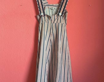 May include: A sleeveless midi dress featuring a vertical striped pattern in navy blue, white, and peach. The dress has wide shoulder straps with a floral trim, a square neckline, and a central front zipper closure with two white buttons at the waist.