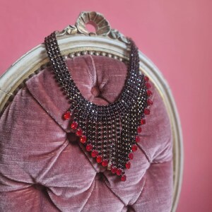 Può includere: Una collana di perline nere con dettagli rossi è appoggiata su una sedia di velluto rosa trapuntata. La collana presenta un design a cascata con file di perline nere e gemme rosse vibranti, creando un contrasto sorprendente con la trama della sedia.