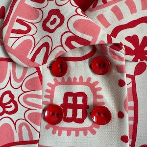 May include: A double-breasted jacket in an off-white fabric featuring a bold, graphic pattern of red and pink abstract shapes and floral motifs. The garment includes a wide collar, a red interior lining, and four large, round, glossy red buttons.