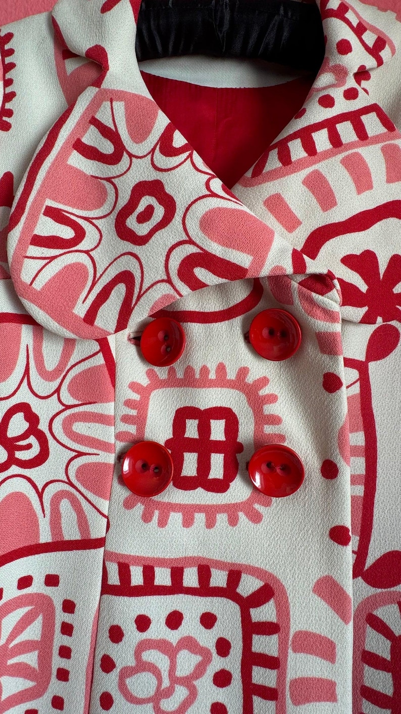 May include: A double-breasted jacket in an off-white fabric featuring a bold, graphic pattern of red and pink abstract shapes and floral motifs. The garment includes a wide collar, a red interior lining, and four large, round, glossy red buttons.