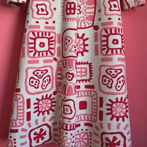May include: A long-sleeved coat featuring an off-white base color with a repeating grid pattern of various geometric shapes and abstract symbols in shades of bright pink and dark red, extending from the shoulders down to the hemline.