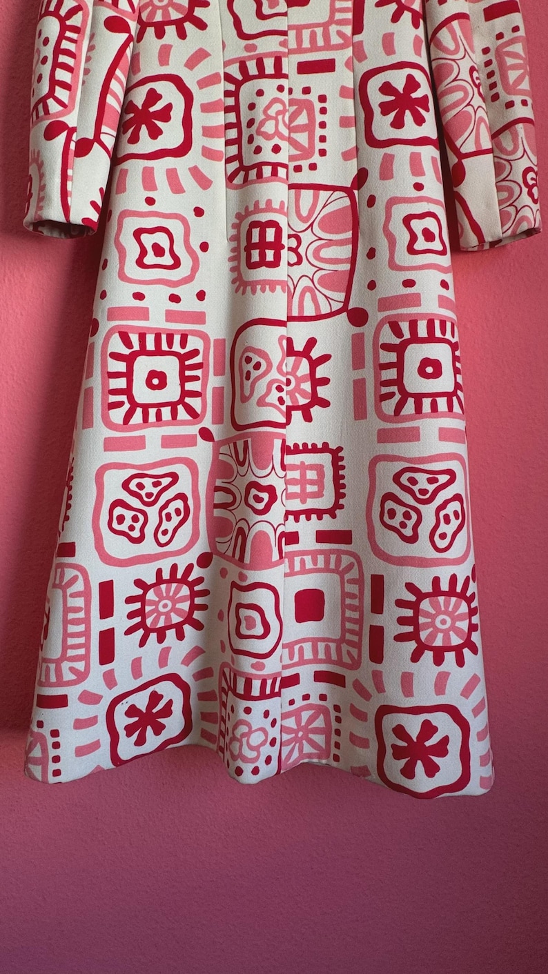 May include: A long-sleeved coat featuring an off-white base color with a repeating grid pattern of various geometric shapes and abstract symbols in shades of bright pink and dark red, extending from the shoulders down to the hemline.