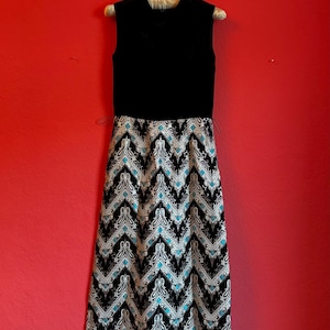 Può includere: Un abito nero senza maniche con gonna fantasia. La gonna presenta un motivo a zig-zag nero, bianco e turchese. L'abito è appeso a una gruccia di legno su sfondo rosso.