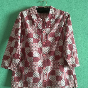 Könnte beinhalten: Ein Hemd mit Knopfleiste und Patchwork-Muster, bestehend aus abwechselnden Quadraten in Altrosa und weißem Stoff mit kleinen blauen Blumenmustern. Das Hemd hat einen spitzen Kragen, Dreiviertelärmel und dazu passende rosafarbene Knöpfe.