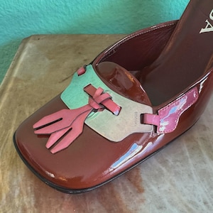 May include: A brown patent leather shoe with a square toe and a high heel. The shoe features a decorative accent with pink and green elements. The brand name is visible on the side of the shoe.