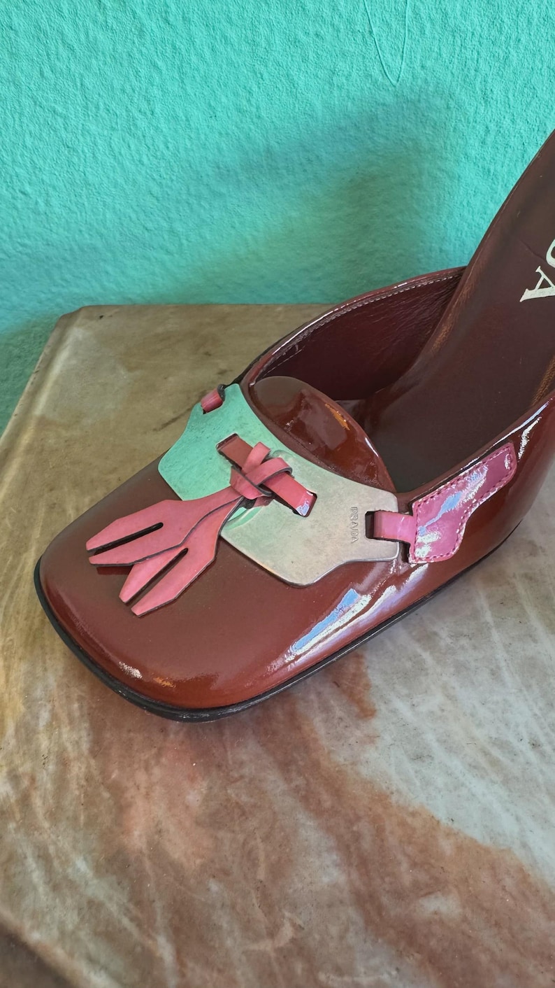 May include: A brown patent leather shoe with a square toe and a high heel. The shoe features a decorative accent with pink and green elements. The brand name is visible on the side of the shoe.