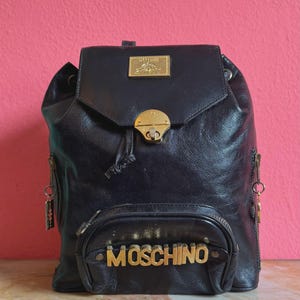 May include: A black leather backpack featuring a gold-tone metal Moschino logo plate on the front pocket. The bag includes a top flap with a gold-tone metal clasp closure, a rectangular brand plaque, and side zippers with hanging metal charms.